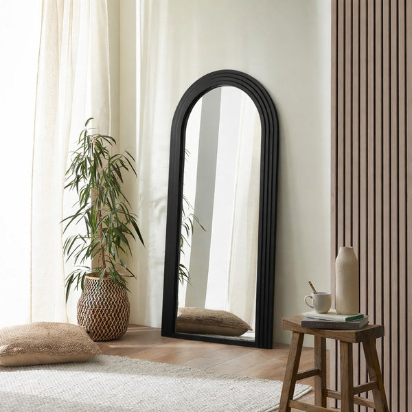 Black arch-shaped mirror in a room with a plant, stool, and cushions.
